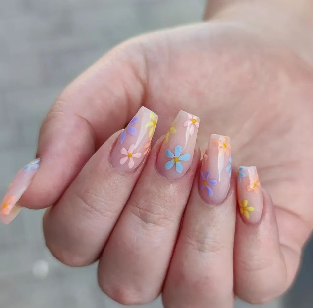 A woman's hand showcasing a glossy nude nails are adorned with delicate flowers in shades of blue, lavender, yellow, and pink