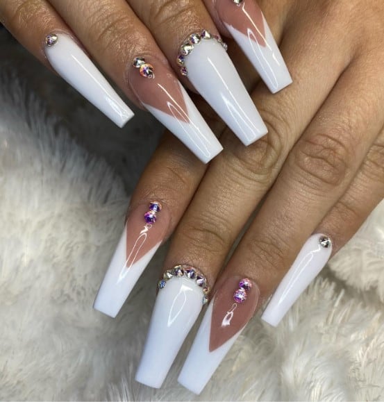 A woman's nails feature classic plain white nails adorned with jewel studs framing the nail base and deep white V-tip coffin nails with a delicate pink base embellished with two vertical gems at the cuticles.