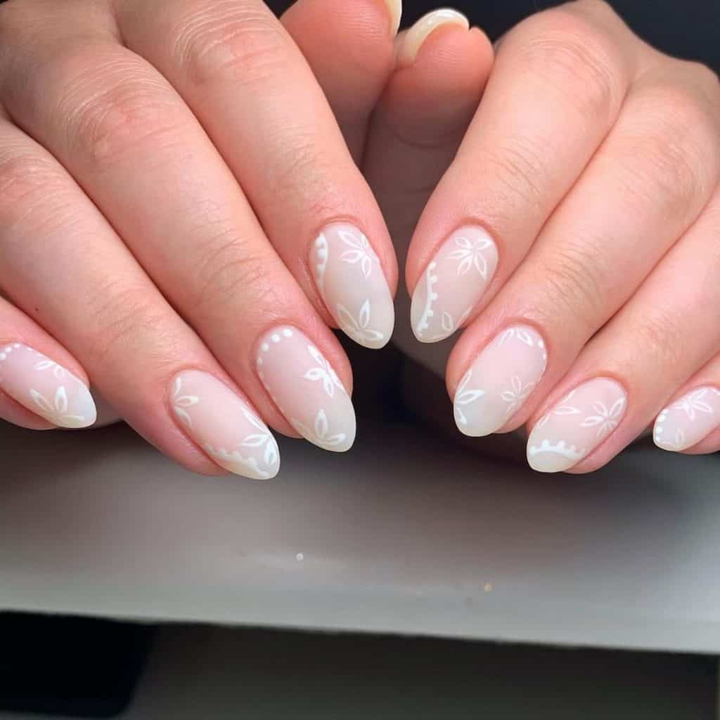 A woman's hands with matte nails in sheer white are painted with intricate patterns of dots, swirls, and flower outlines in crisp white