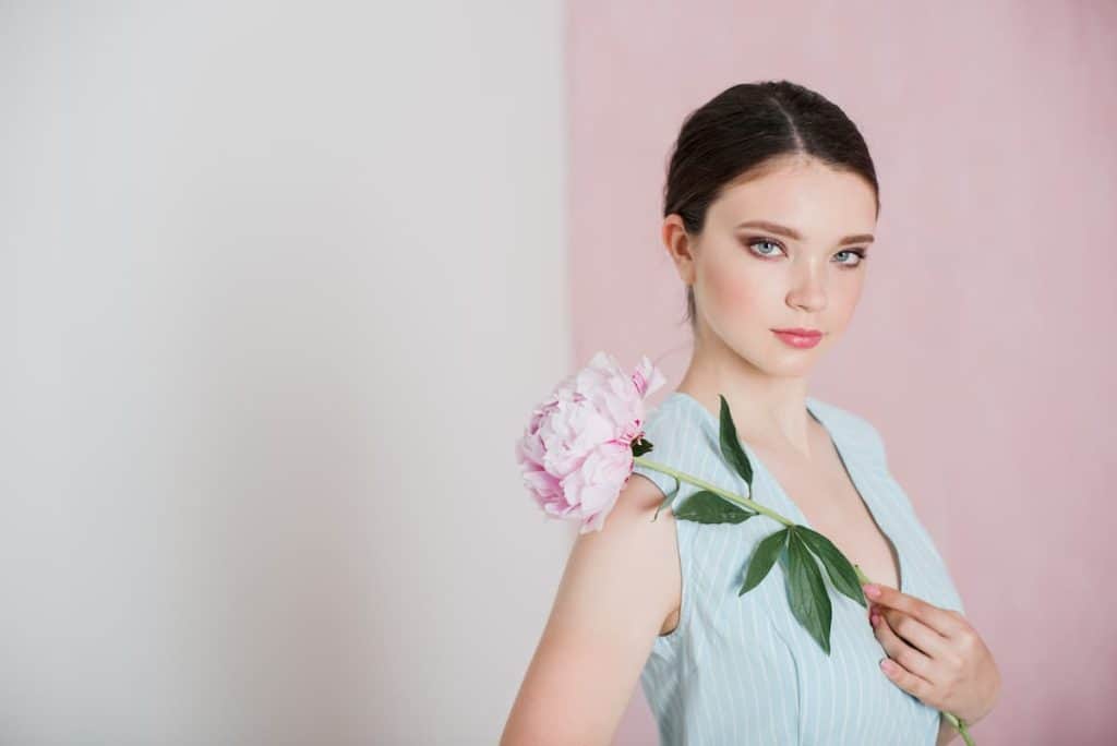A young beautiful woman in a blue striped dress with gathered hair is standing holding a peony in her hands.
