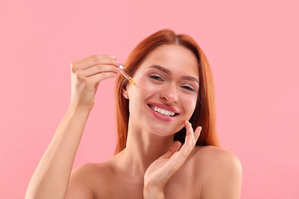 Beautiful young woman applying cosmetic serum onto her face on pink background