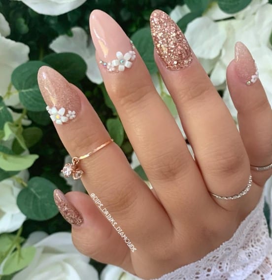 Glimmering with fine glitter, two nude almond nails bloom with 3D white flowers while their rose gold glitter twins shimmer. 