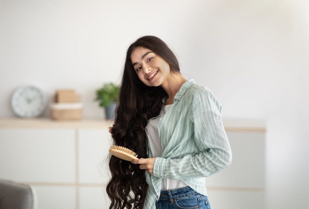 Portrait of lovely Indian lady brushing her wavy long hair, using wooden brush indoors. 
