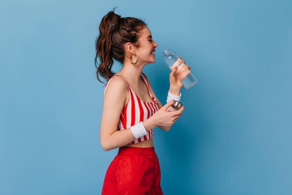 Young girl in red striped outfit laughs and drinks water from bottle on isolated background
