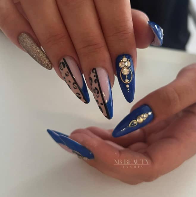 A woman's  long, blue almond nails combine several designs, including unique blue French tips on a pale pink base, a black line down the middle with a glittery cheetah print, full blue nails studded with jewels, and pinky nails dipped in glittering gold