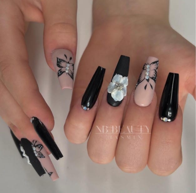 A woman's black coffin nails have multiple designs: some have rhinestones close to the cuticles, while some have a prominent white 3D flower