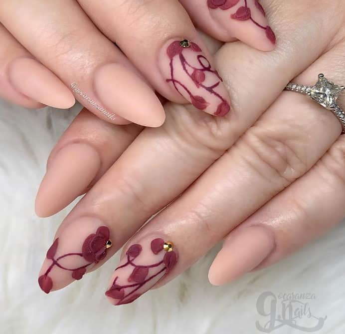 These nude peach almond nails in matte are a canvas for creativity, with accent nails showcasing embossed dark red petal art and rhinestones.