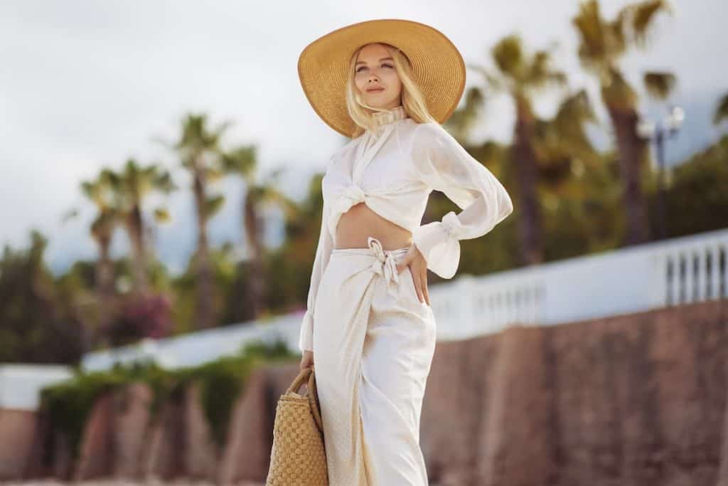 A blonde woman in a hat and white pants standing on the beach.