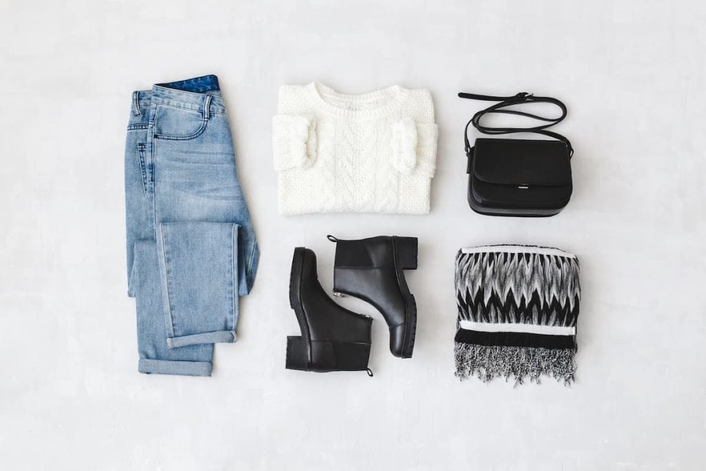 Blue jeans, white knitted sweater, small black cross body bag, leather ankle boots and striped scarf on grey background.