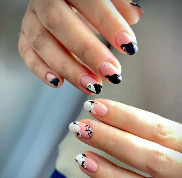 A woman's nails with short light pink oval claws, one hand tipped white with black webs and stars, and the other tipped black with white ghostly webs and shining stars