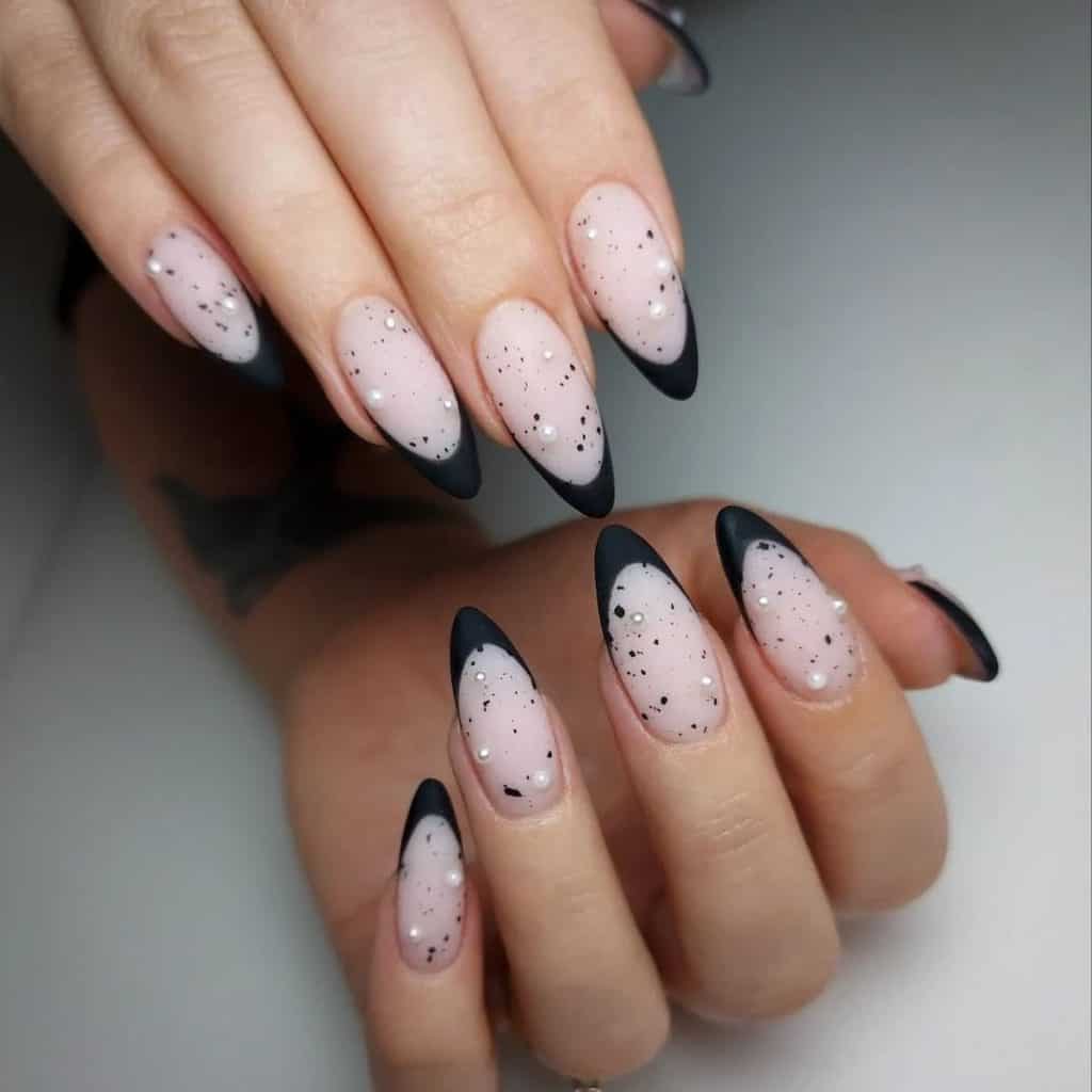 Sheer matte nude pink nails with bold black French tips are sprinkled with black speckles and adorned with tiny pearl studs