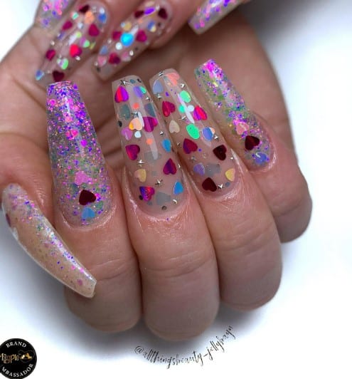 A woman's nails with translucent coffin nails decorated with a confetti of magenta, purple, blue, and iridescent chunky glitter, along with some heart-shaped ones