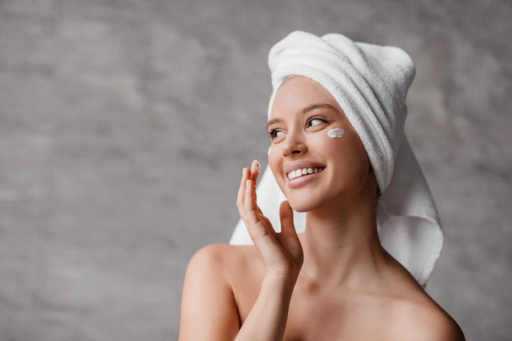 Sexy lady applying moisturizer cream under eyes, doing facial skin care, standing in bathroom, looking aside and smiling, free space