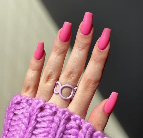 A woman wearing a purple sweater with pink nails shaped into coffin style and a pink ring.