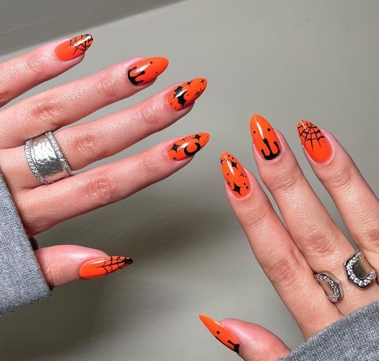 A woman's nails with bright orange and decorated with black designs such as spiderwebs at the tips, moons, twinkling stars, and dripping blood
