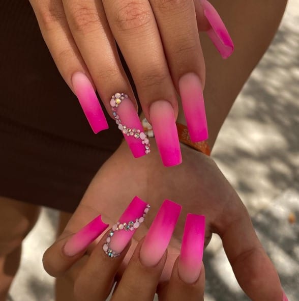 A woman's pink matte nails with rhinestones feature mostly ombré ones, save for one that’s crowned with swirls of rhinestones and pearl studs for a touch of opulent charm