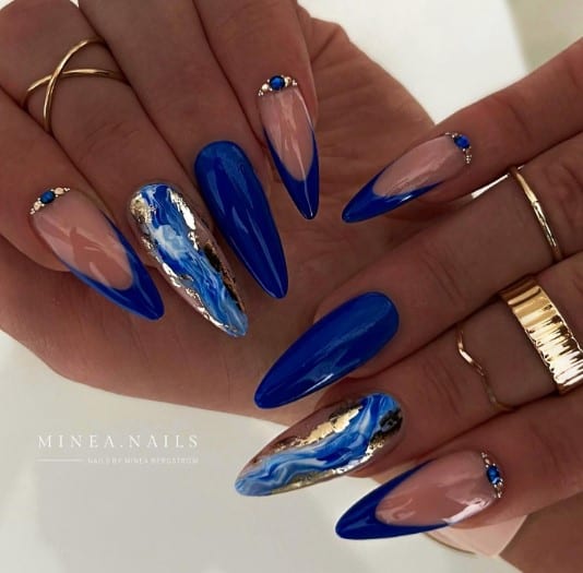 A woman with blue almond nails pair a nude peach base with royal blue French tips and are embellished with diamond studs and sapphire stones