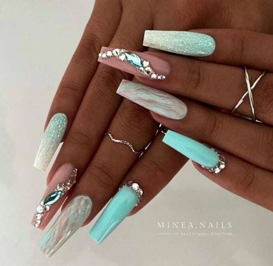 Feature glossy aqua nails with sparkling rhinestones, a shimmering blue and white ombré, clear nails with white and blue swirls, and a nail with an S-shaped pattern of rhinestones on a nude base.