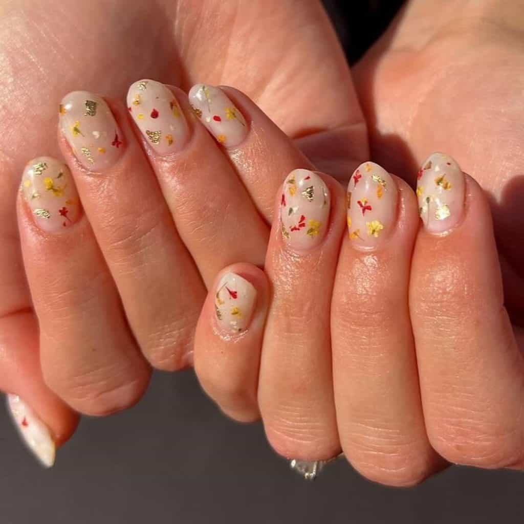 A woman's short and round nails in a nude white embrace adorned with flecks of gold foil and tiny blossoms in yellow and red