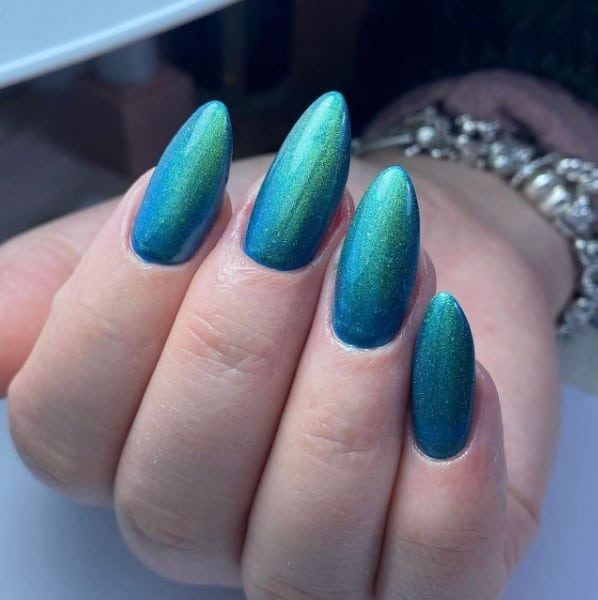 A woman's long blue almond nails, boasting cosmic allure as green glittery dust gracefully cascades on each nail