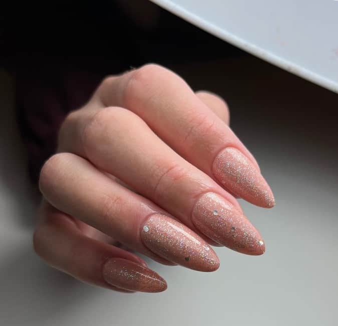 Glittery nude almond nails twinkle with flecks of silver, casting a shimmering glow with every gesture — a dash of sparkle for your daily dazzle.