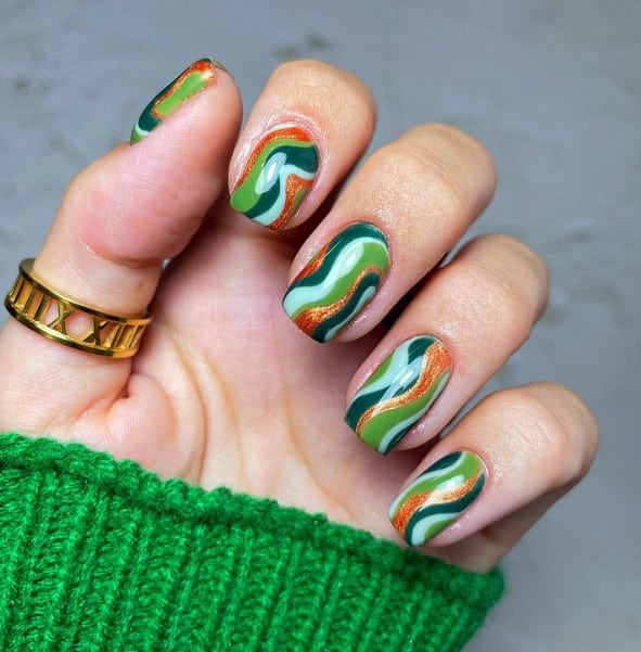 a woman's nails with playful design that blends swirls of various green shades ranging from light to dark with a glittery bronze for contrast