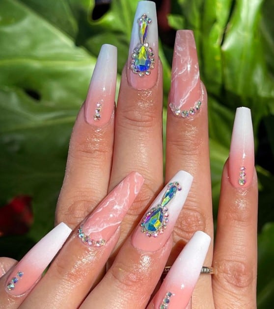 A woman's coffin nails with three sporting a nude-to-white ombré adorned with crystals and rhinestones.