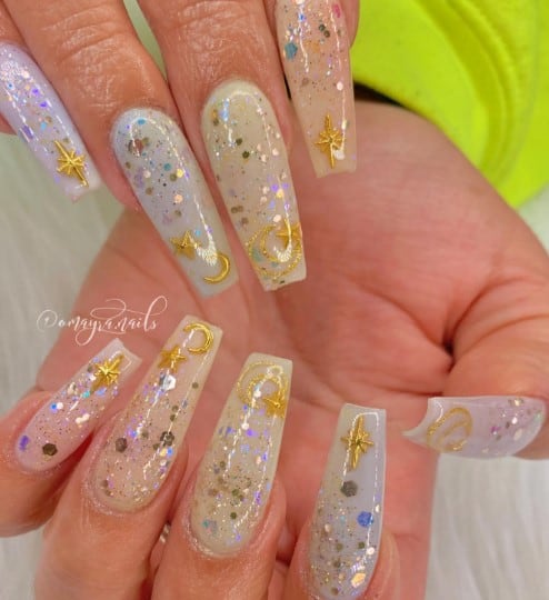 A woman's nails that boast a sheer white base, sparkling with chunky glitters and golden celestial motifs, including North Stars, crescent moons, and twinkling stars at the tips