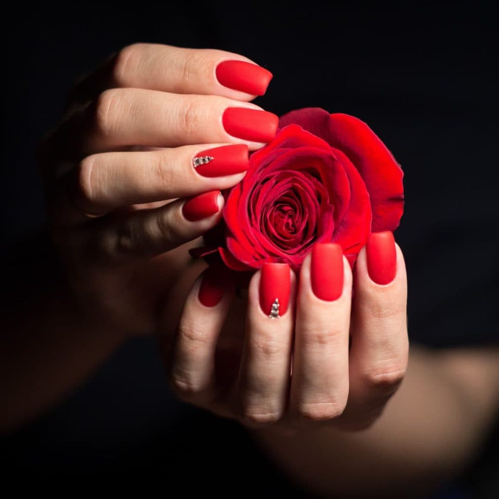 A woman's matte red nails, each accent nail tastefully embellished with a rhinestone near the cuticles