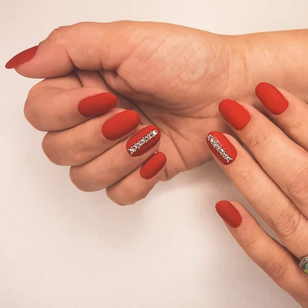 This matte red nail design features an accent nail that showcases a vertical line of rhinestones and silver round studs at the center.