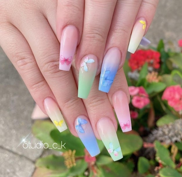 A woman's hand each nail featuring a butterfly atop different ombré nails transitioning from a nude base to either yellow, blue, mint green, or pink