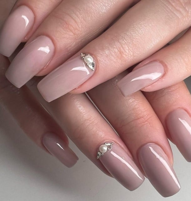A woman's hands with glossy nude pink square nails, complemented with accent nails that flaunt pearls and tiny gems at the cuticles
