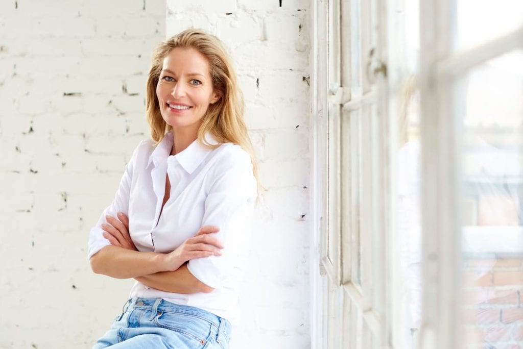 A smiling woman leaning against a window while wearing white buttondown top and a blue jeans