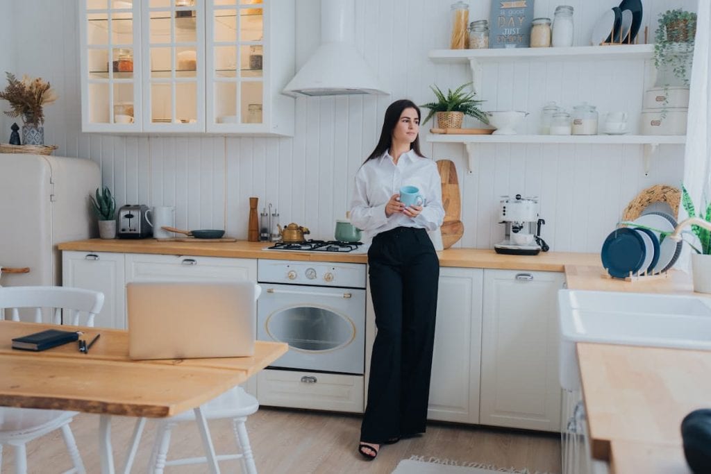 Thoughtful brunette young woman in white shirt and black pants holding cup of coffee smiles wide looks aside stands at kitchen.