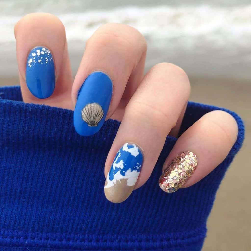 A sprinkle of silver glitter flakes at the cuticles mimics a gentle ocean spray, while sandy clamshell art graces another nail. The third captures the serene feel of a sandy seashore with seafoam art.