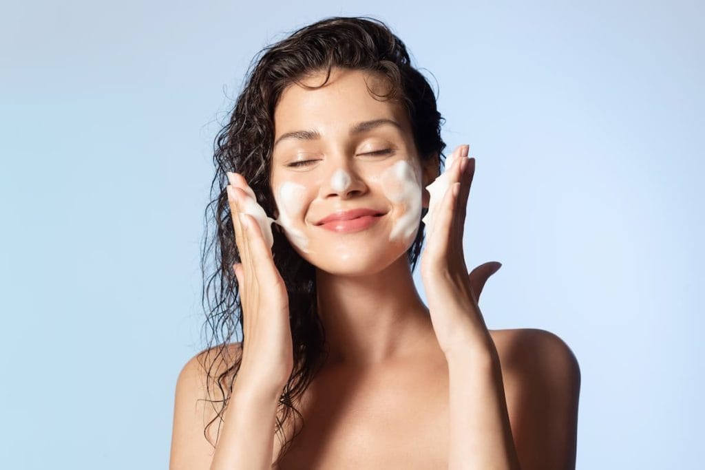Satisfied girl with bare shoulders applying cleansing beauty product on cheeks closed eyes.