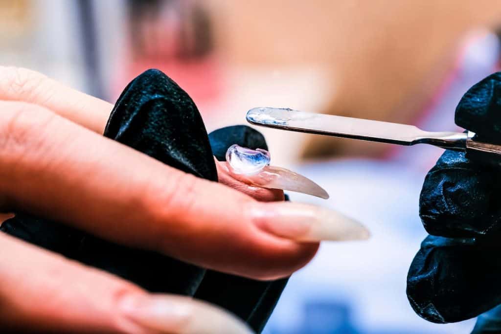 The manicure master applies a base layer of Poly-gel to the nail with a metal spatula. The brush distributes the gel over the entire surface of the nail
